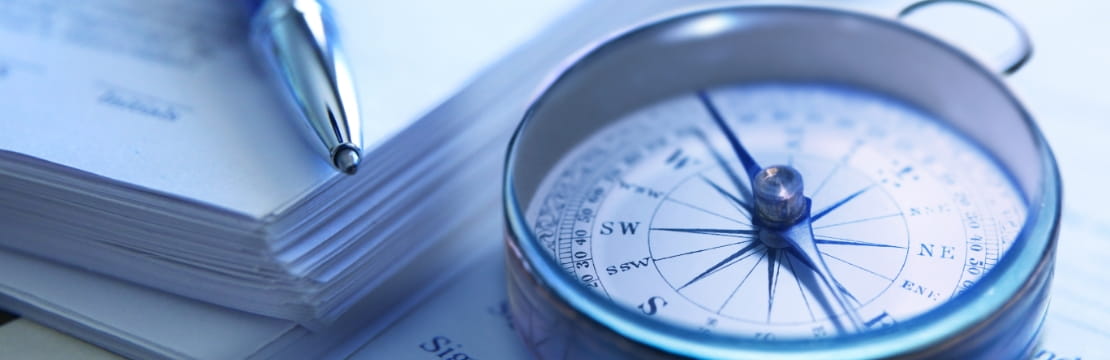 A close-up of a metal compass resting on documents beside a pen.